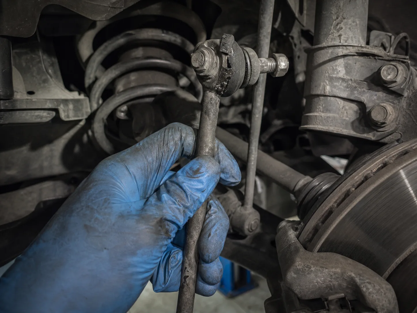 A gloved hand rocking a worn stabiliser link on a raised car, showing visible play at the ball socket